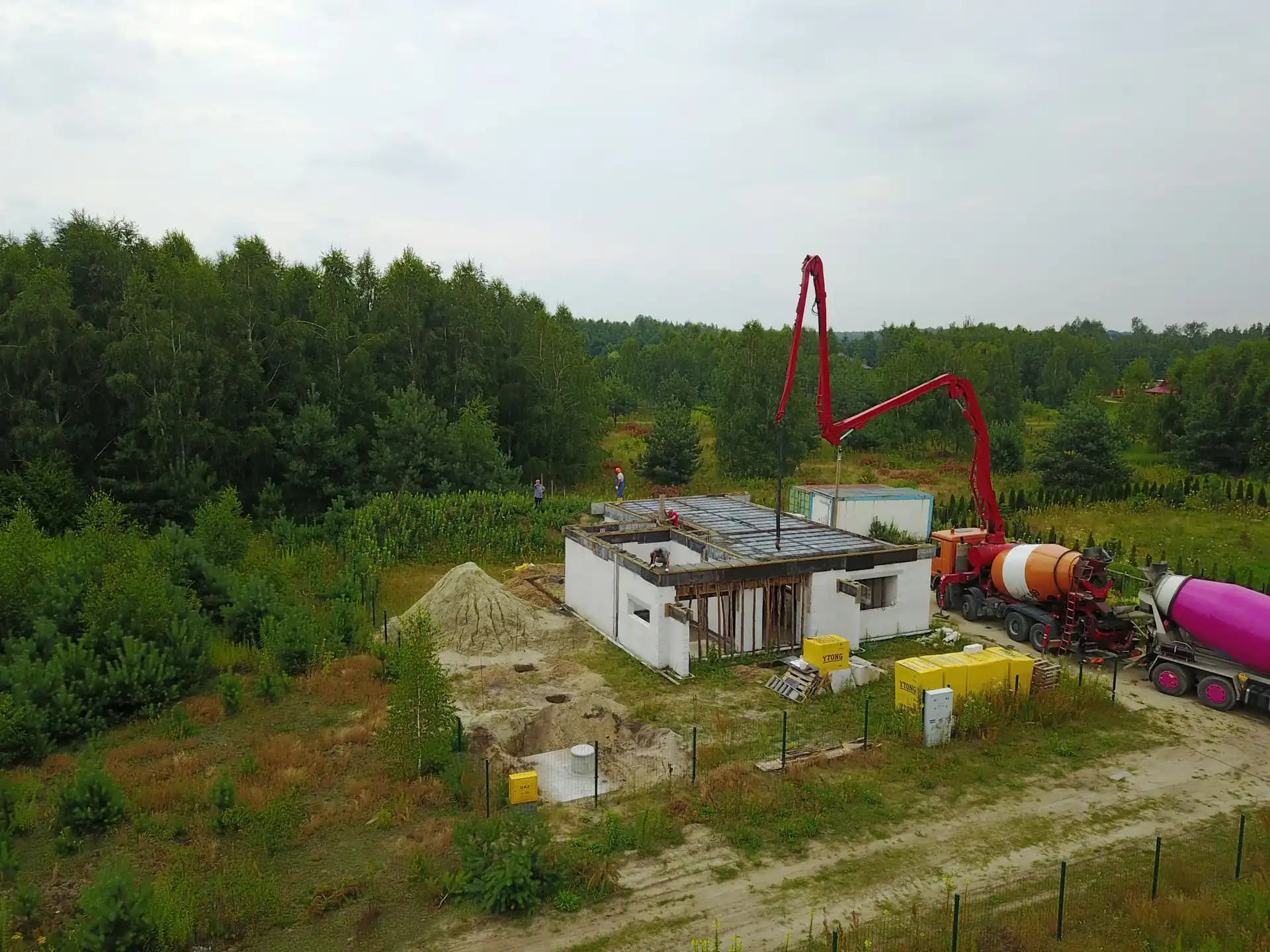 Pompa do betonu z długim ramieniem nad budowanym domem jednorodzinnym, betoniarka i robotnicy w trakcie zalewania stropu, drone shot z perspektywy lotniczej